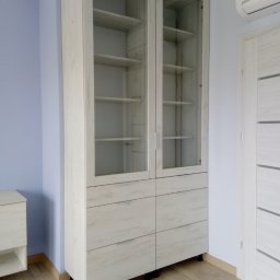 Edelgran.pl - Biała szafa z przeszklonymi drzwiami i szufladami, widoczna na tle błękitnej ściany i drewnianej podłogi.