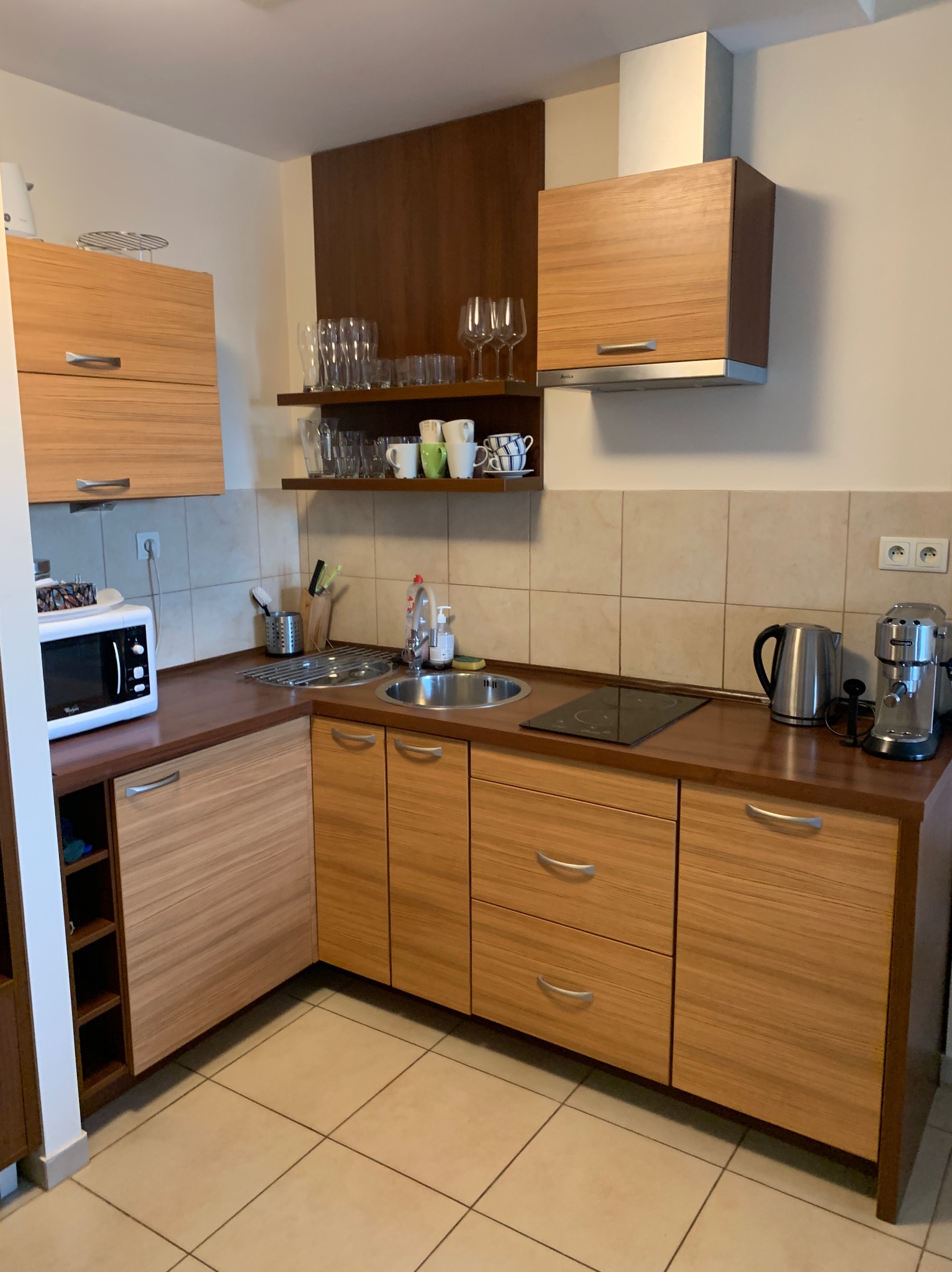 Jasna kuchnia z drewnianymi frontami, wyposażona w kuchenkę indukcyjną, zlew, mikrofalówkę i ekspres do kawy, z otwartą półką z naczyniami i akcesoriami kuchennymi.