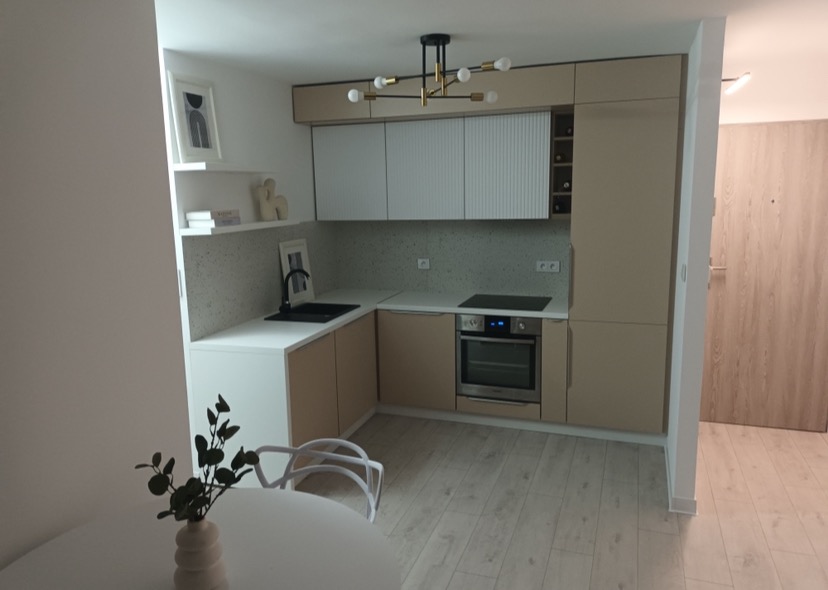 Nowoczesna, beżowo-biała kuchnia z elementami drewna, oświetlona designerską lampą sufitową, widok z perspektywy jadalni z białym stołem i krzesłami.