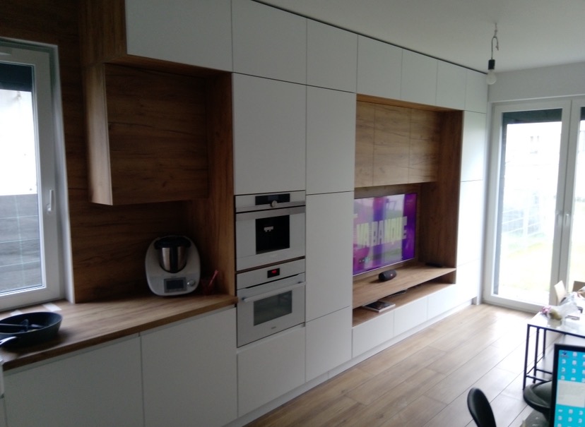 Nowoczesna zabudowa kuchenna z białymi frontami i elementami z drewna, z wbudowanymi piekarnikami i przestrzenią na telewizor w salonie.