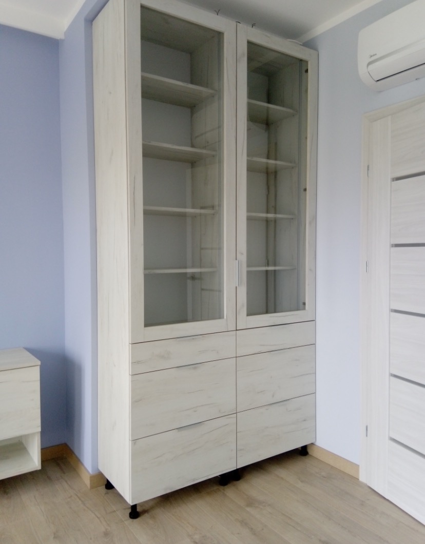Biała szafa z przeszklonymi drzwiami i szufladami, widoczna na tle błękitnej ściany i drewnianej podłogi.