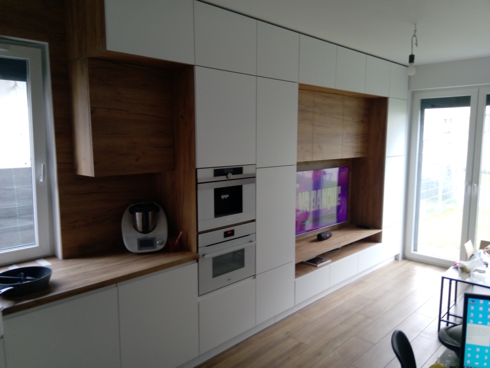Nowoczesna zabudowa kuchenna z białymi frontami i drewnianymi wstawkami, obejmująca piekarnik, ekspres do kawy, półki i blat z robotem kuchennym, widok na salon z telewizorem.