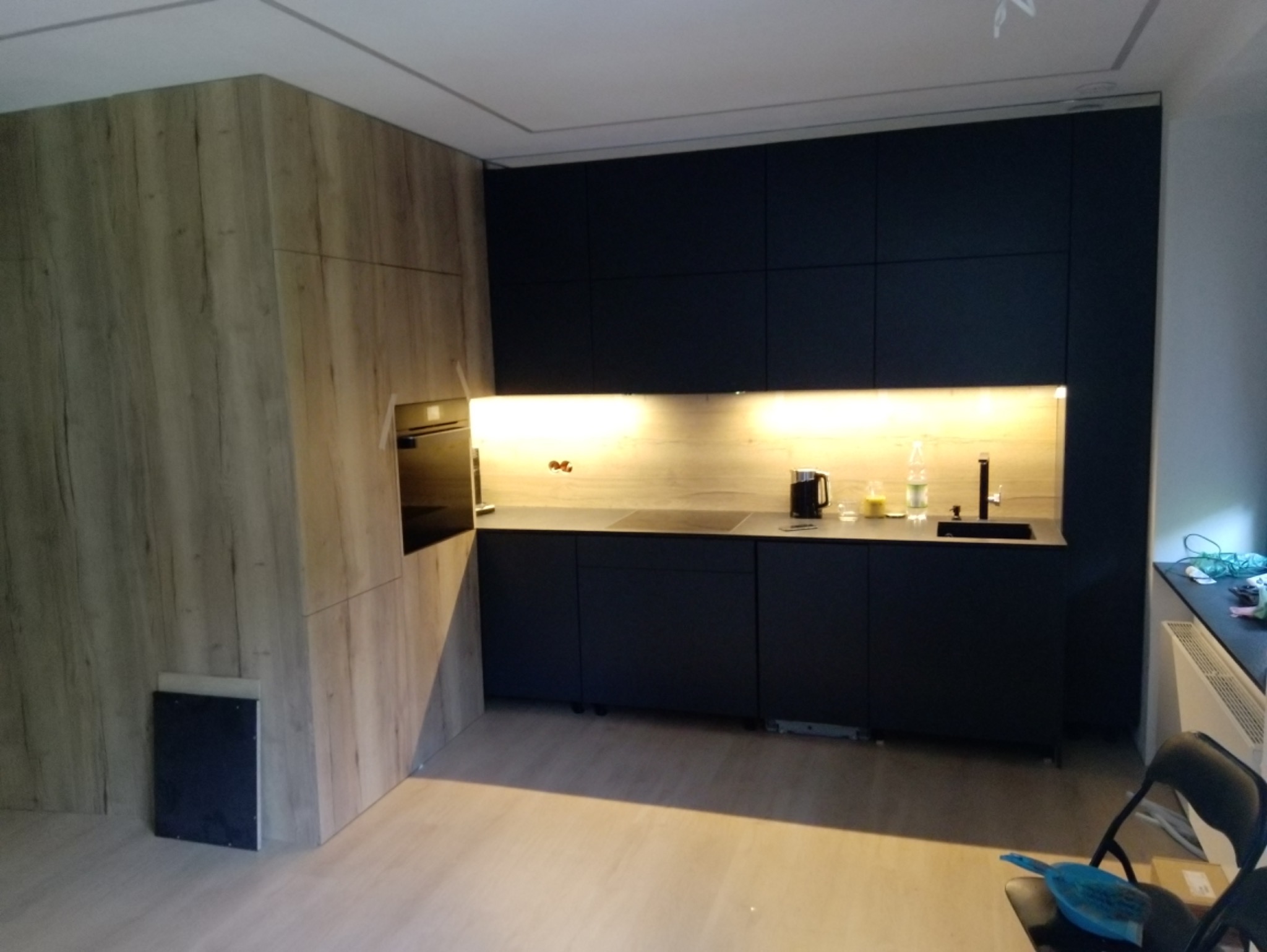 Nowoczesna kuchnia z zabudową w odcieniach drewna i grafitu, podświetlane blaty, widoczne akcesoria kuchenne i elementy wykończeniowe.