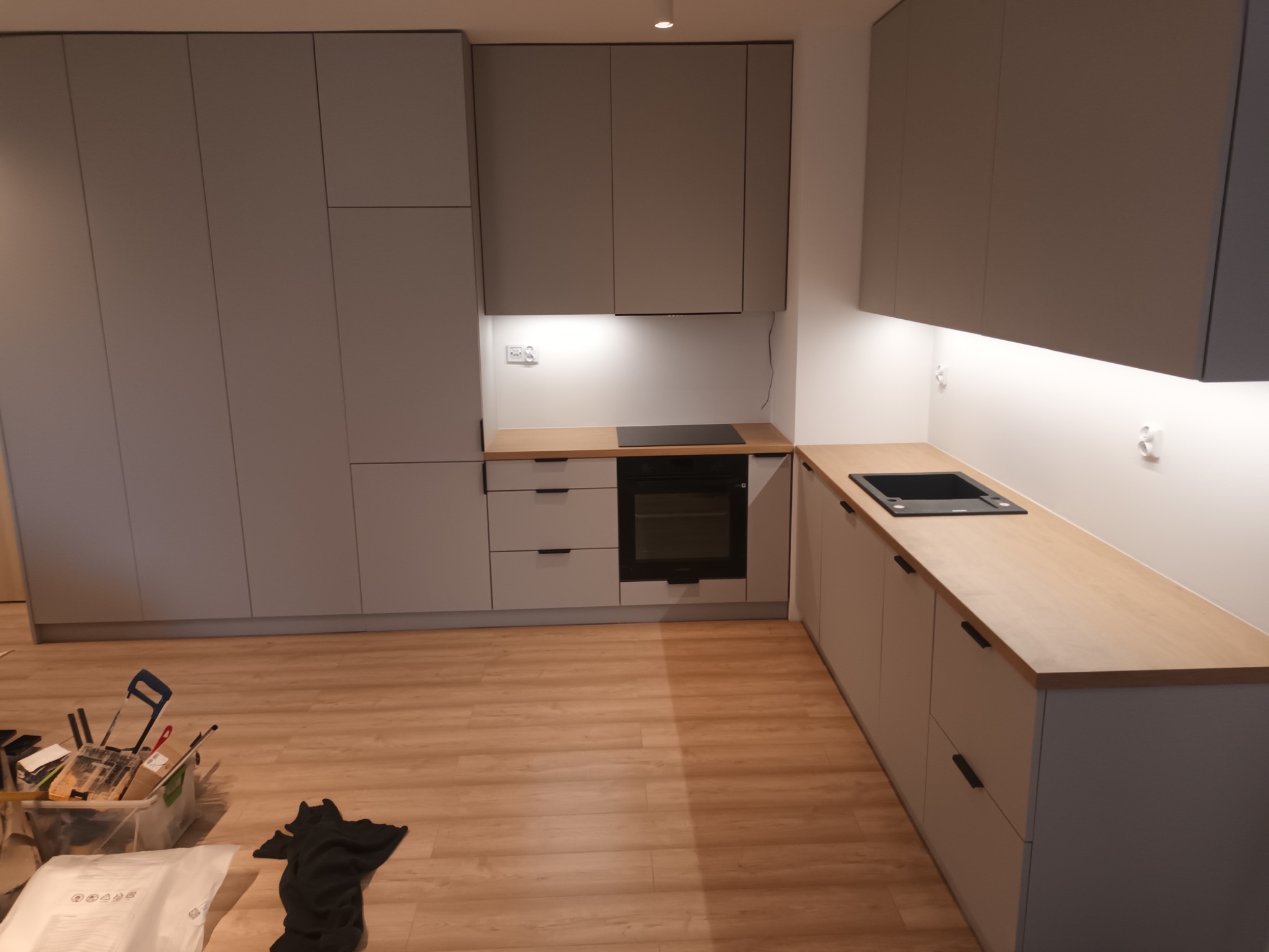 Szare szafki kuchenne na wymiar z drewnianym blatem, czarnym zlewem i płytą indukcyjną, widoczny bałagan po remoncie.