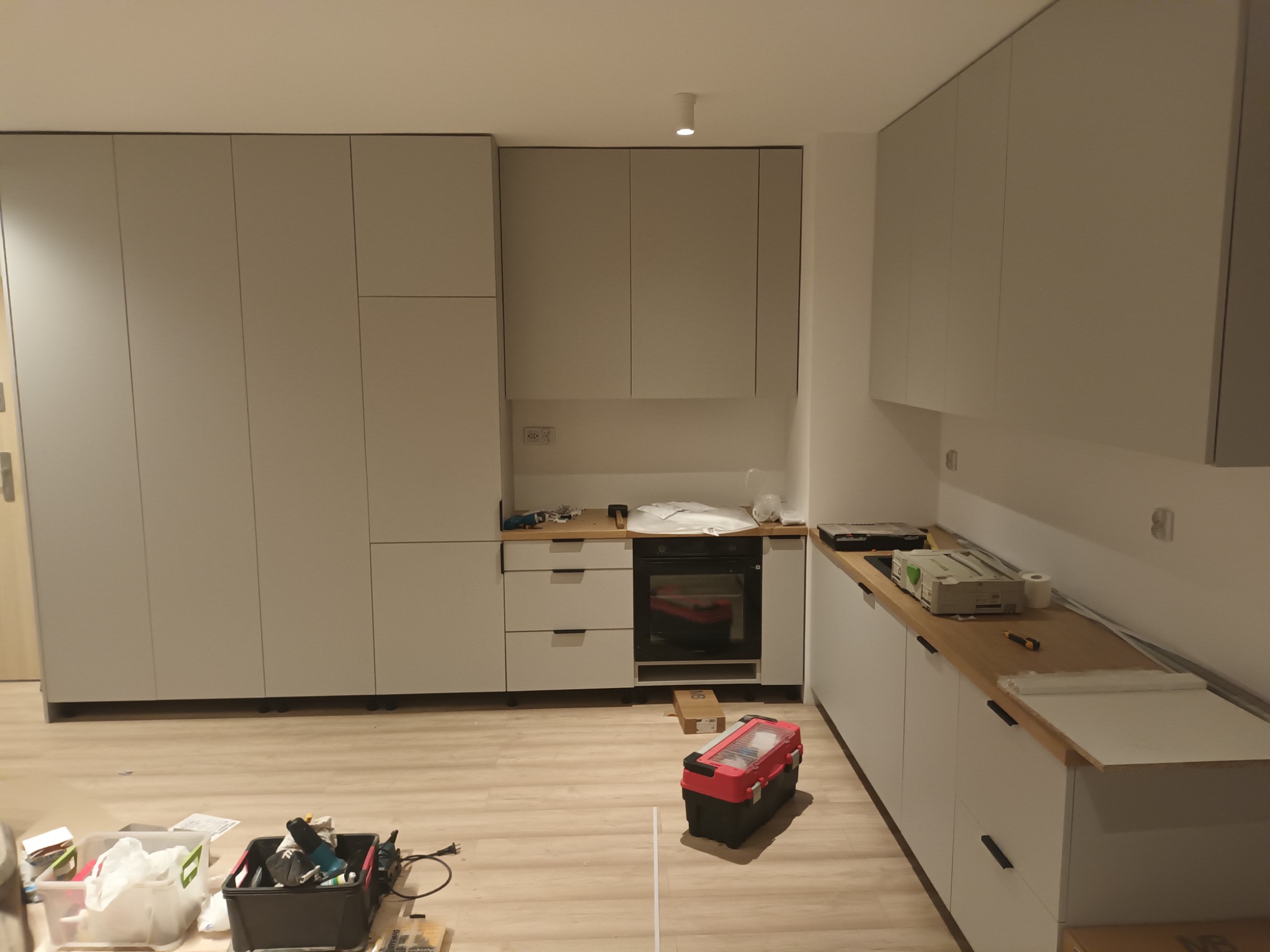 Montaż szarych szafek kuchennych z drewnianym blatem, widoczne narzędzia i skrzynka narzędziowa na podłodze, oświetlenie sufitowe.