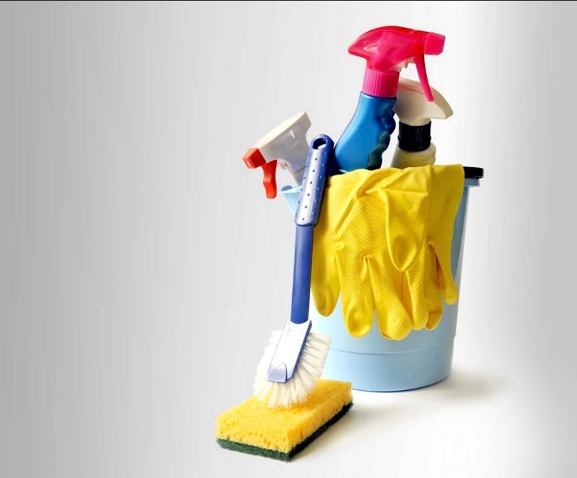 Niebieskie wiadro z żółtymi rękawicami, butelkami ze spryskiwaczem, szczotką i gąbką na szarym tle. Zestaw do sprzątania.