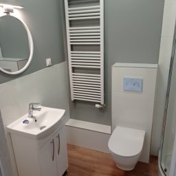 Damian Grzelak - Nowoczesna łazienka z białymi płytkami i szarą ścianą, wyposażona w wiszącą toaletę z niebieskim przyciskiem spłukiwania, umywalkę z szafką oraz drabinkowy grzejnik.