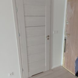 Damian Grzelak - Zamontowane białe drzwi wewnętrzne z poziomymi panelami, widoczne gniazdko elektryczne na ścianie, podłoga laminowana, w tle drzwi zabezpieczone folią malarską.