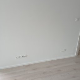 Damian Grzelak - Świeżo pomalowana, biała ściana z zamontowanymi gniazdkami elektrycznymi i wykończoną listwą przypodłogową, jasna podłoga panelowa.