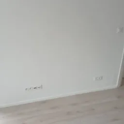 Świeżo pomalowana, biała ściana z zamontowanymi gniazdkami elektrycznymi i wykończoną listwą przypodłogową, jasna podłoga panelowa.