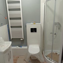 Damian Grzelak - Nowoczesna łazienka w trakcie wykańczania: biała kabina prysznicowa, podwieszana toaleta zabezpieczona folią, grzejnik drabinkowy na szarej ścianie, widoczne narzędzia i niedokończona podłoga.