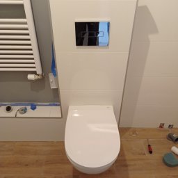 Damian Grzelak - Podwieszana toaleta z białą zabudową i czarnym panelem spłukującym, widoczne narzędzia i akcesoria na podłodze z płytek imitujących drewno, grzejnik drabinkowy po lewej stronie, etap montażu.