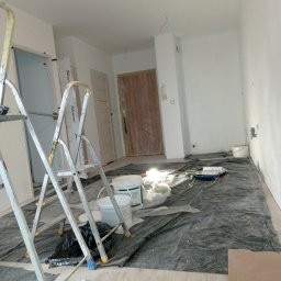 Damian Grzelak - Pokój w trakcie odświeżania: zabezpieczona podłoga, drabiny malarskie, wiadra z farbą, wałek i kuweta malarska, białe ściany po malowaniu.
