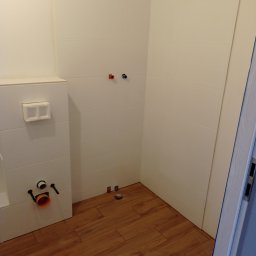 Damian Grzelak - Łazienka w trakcie przygotowania do montażu armatury: białe płytki na ścianach, drewnopodobna podłoga, widoczne przyłącza wodne i odpływowe, częściowo zamontowany stelaż podtynkowy WC.