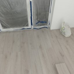Damian Grzelak - Podłoga z paneli w odcieniach szarości, zabezpieczone folią okna i biała ściana podczas prac wykończeniowych, widoczny worek z klejem