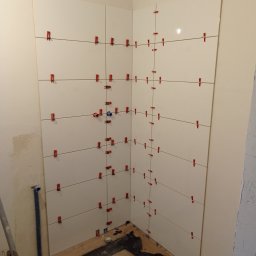 Damian Grzelak - Świeżo ułożone, białe płytki ścienne w rogu łazienki, z czerwonymi klipsami poziomującymi i sznurkami, widoczne rury i fragment podłogi zabezpieczonej folią.