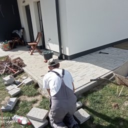 Damian Grzelak - Pracownik w trakcie układania jasnoszarej kostki brukowej na posesji domu jednorodzinnego. Widoczny fragment ułożonej nawierzchni i przygotowane materiały.