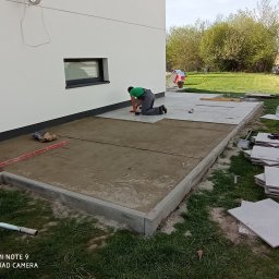 Damian Grzelak - Układanie płyt tarasowych na zewnątrz budynku. Widoczny pracownik na kolanach, poziomica, płyty i przygotowane podłoże. Częściowo ułożona nawierzchnia.