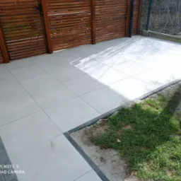 Jasnoszara, kwadratowa płytka tarasowa z ciemnoszarą obwódką, ułożona przy drewnianej konstrukcji w ogrodzie. Część trawnika widoczna w prawym dolnym rogu.