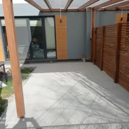 Wyłożony jasnymi płytami taras z drewnianą pergolą, przylegający do szarego budynku z drewnianymi elementami. Widoczny fragment drewnianego ażurowego ogrodzenia.