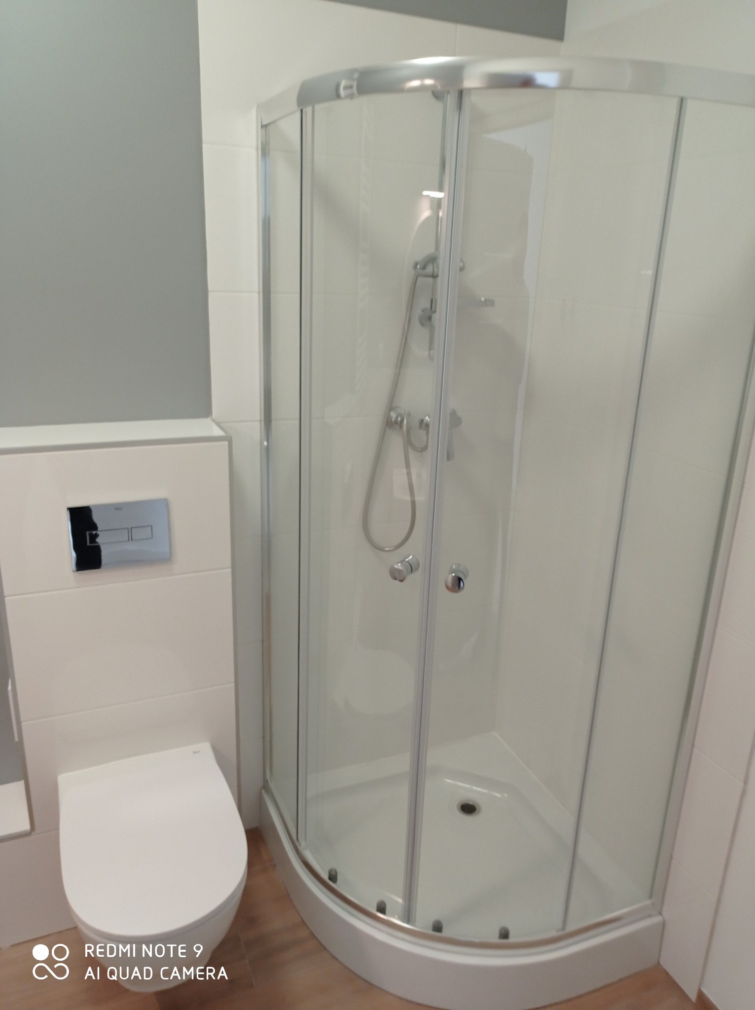 Nowoczesna łazienka z kabiną prysznicową narożną z przezroczystego szkła i podwieszaną toaletą z chromowanym przyciskiem spłukującym, wykończona w bieli i szarości.