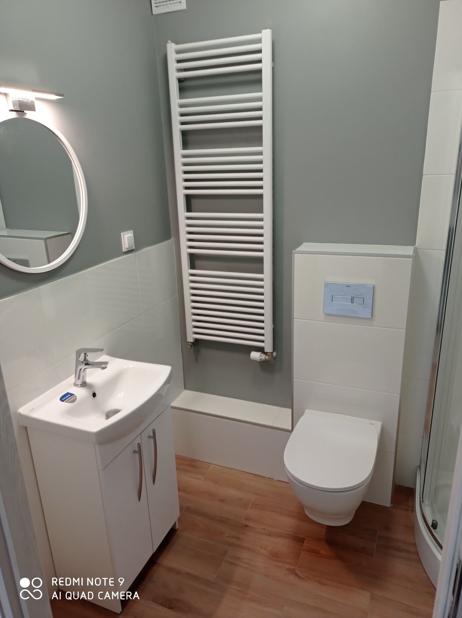 Nowoczesna łazienka z białymi płytkami i szarą ścianą, wyposażona w wiszącą toaletę z niebieskim przyciskiem spłukiwania, umywalkę z szafką oraz drabinkowy grzejnik.