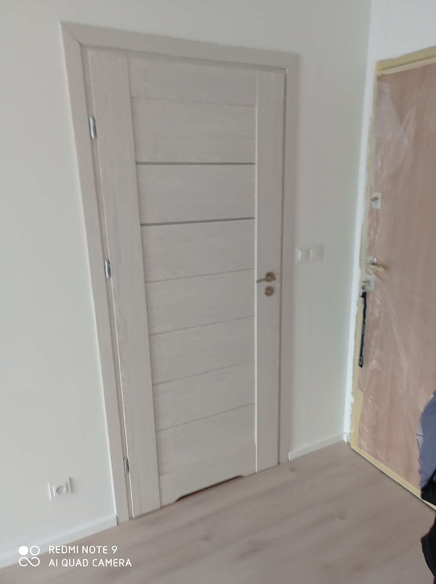 Zamontowane białe drzwi wewnętrzne z poziomymi panelami, widoczne gniazdko elektryczne na ścianie, podłoga laminowana, w tle drzwi zabezpieczone folią malarską.