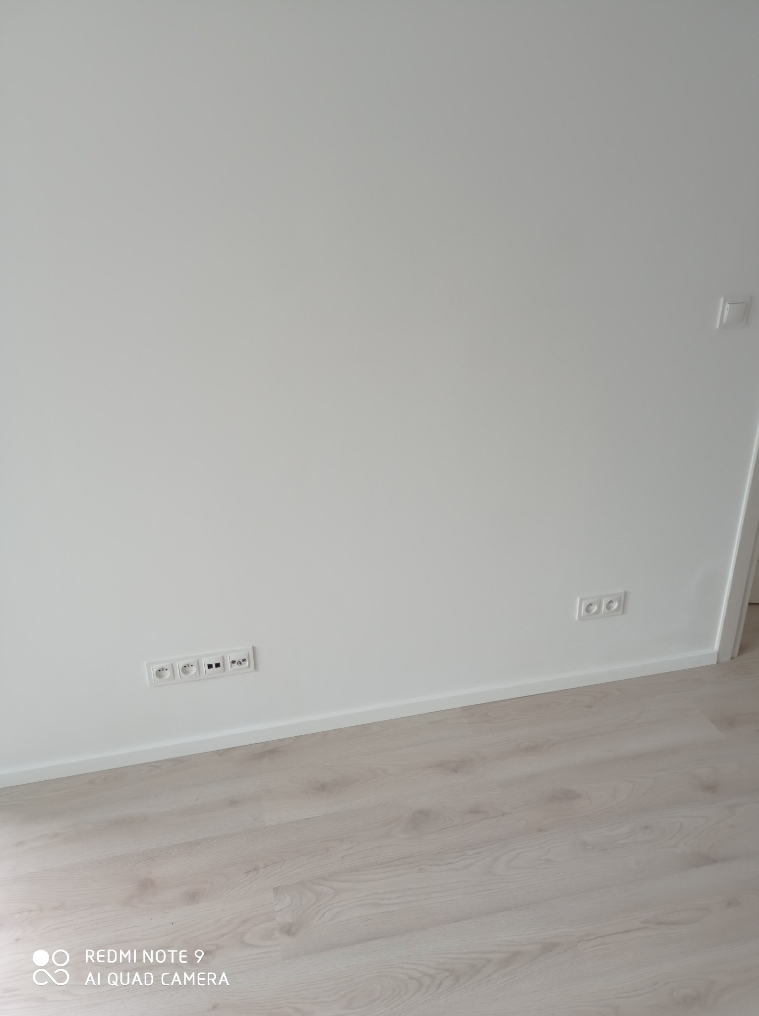 Świeżo pomalowana, biała ściana z zamontowanymi gniazdkami elektrycznymi i wykończoną listwą przypodłogową, jasna podłoga panelowa.