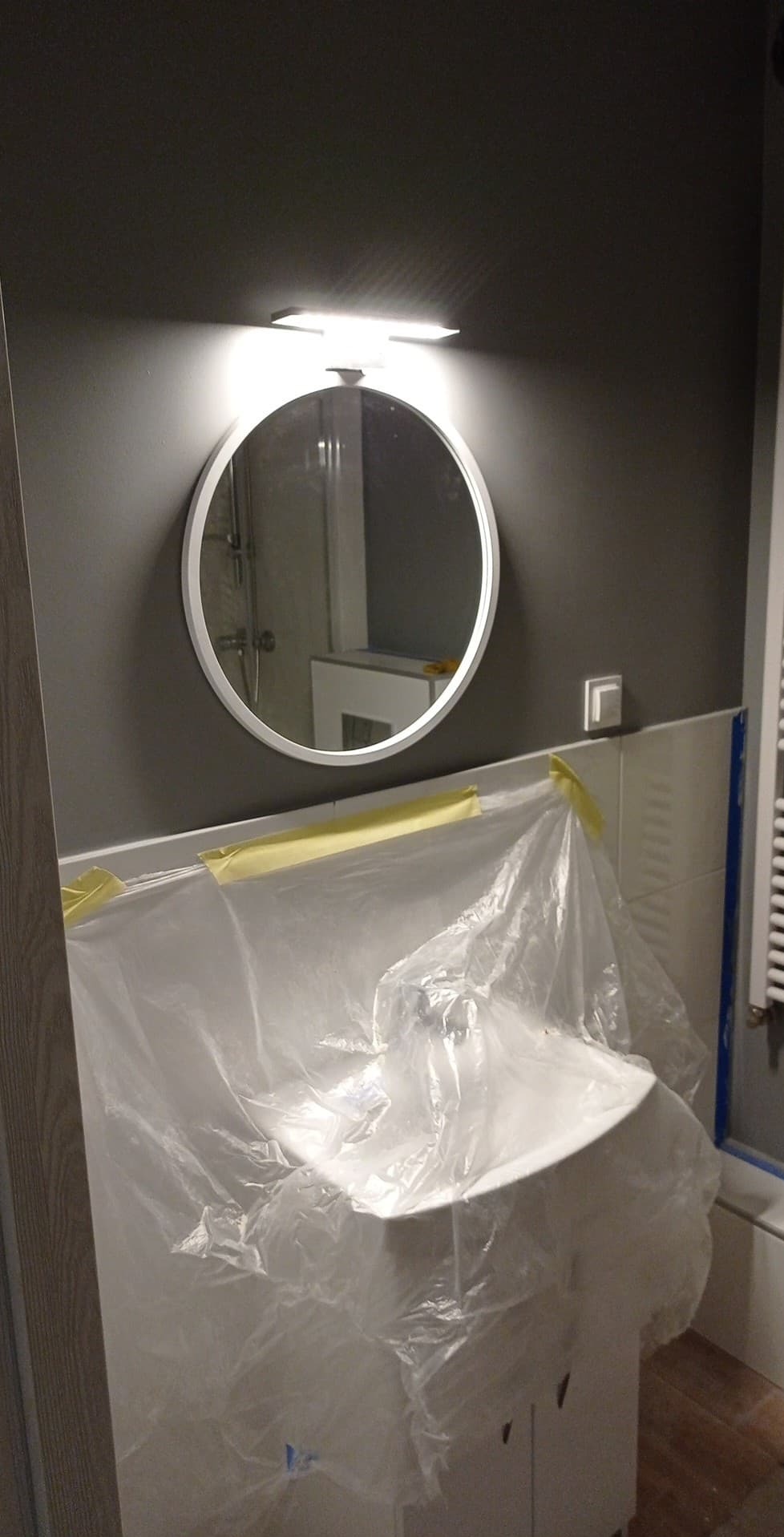 Umywalka z szafką zabezpieczona folią malarską w trakcie prac w łazience w Żyrardowie, okrągłe lustro z podświetleniem LED na szarej ścianie.