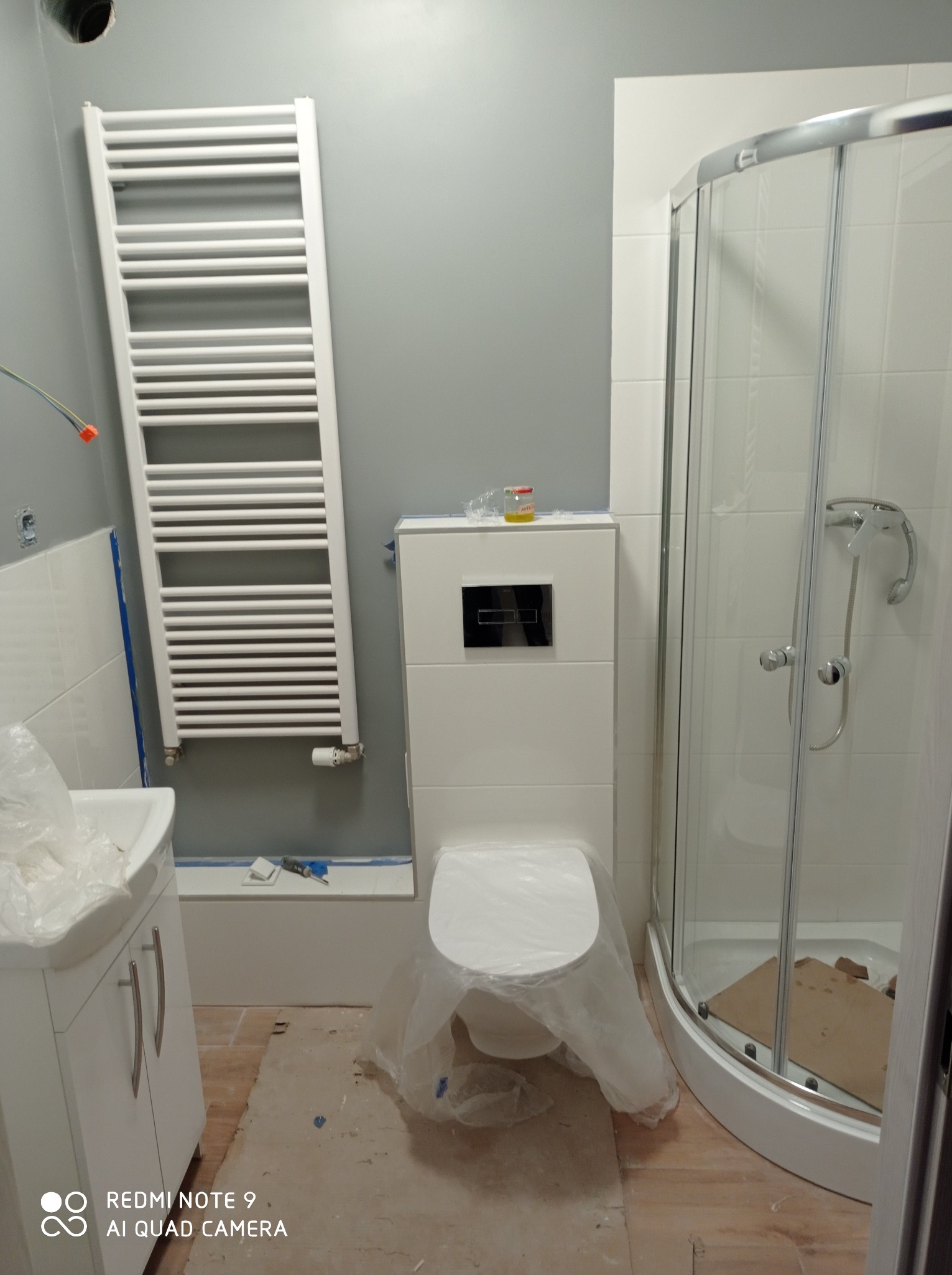 Nowoczesna łazienka w trakcie wykańczania: biała kabina prysznicowa, podwieszana toaleta zabezpieczona folią, grzejnik drabinkowy na szarej ścianie, widoczne narzędzia i niedokończona podłoga.