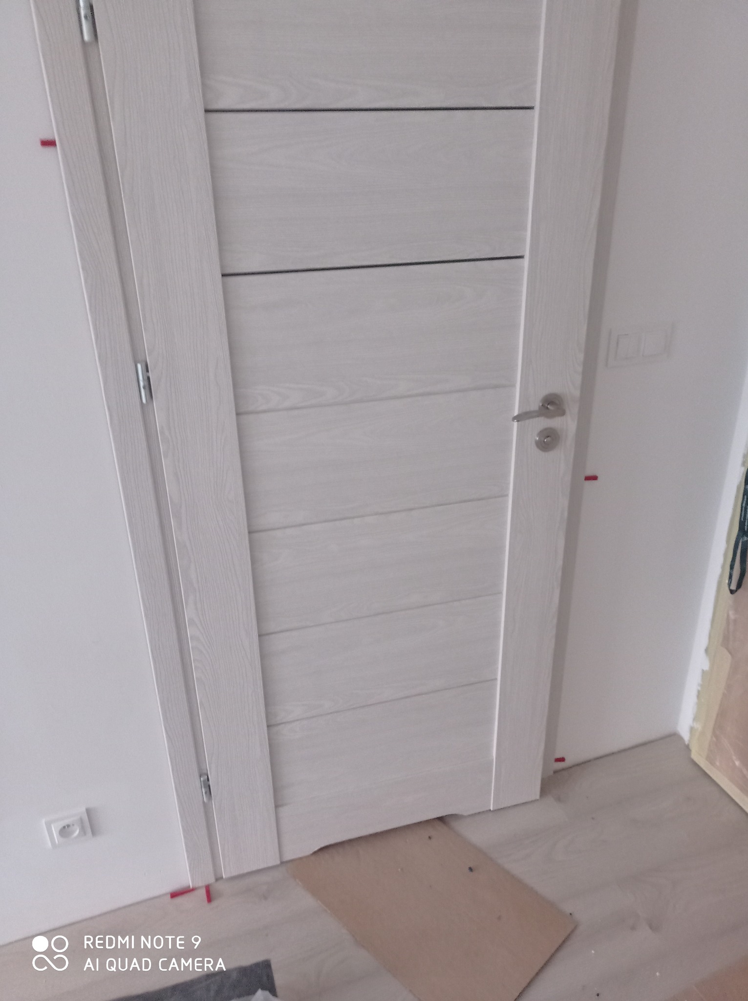 Zamknięte, białe drzwi wewnętrzne z poziomymi żłobieniami, zamontowane w białej ścianie, widoczne fragmenty drewnianej podłogi i listwy przypodłogowe.