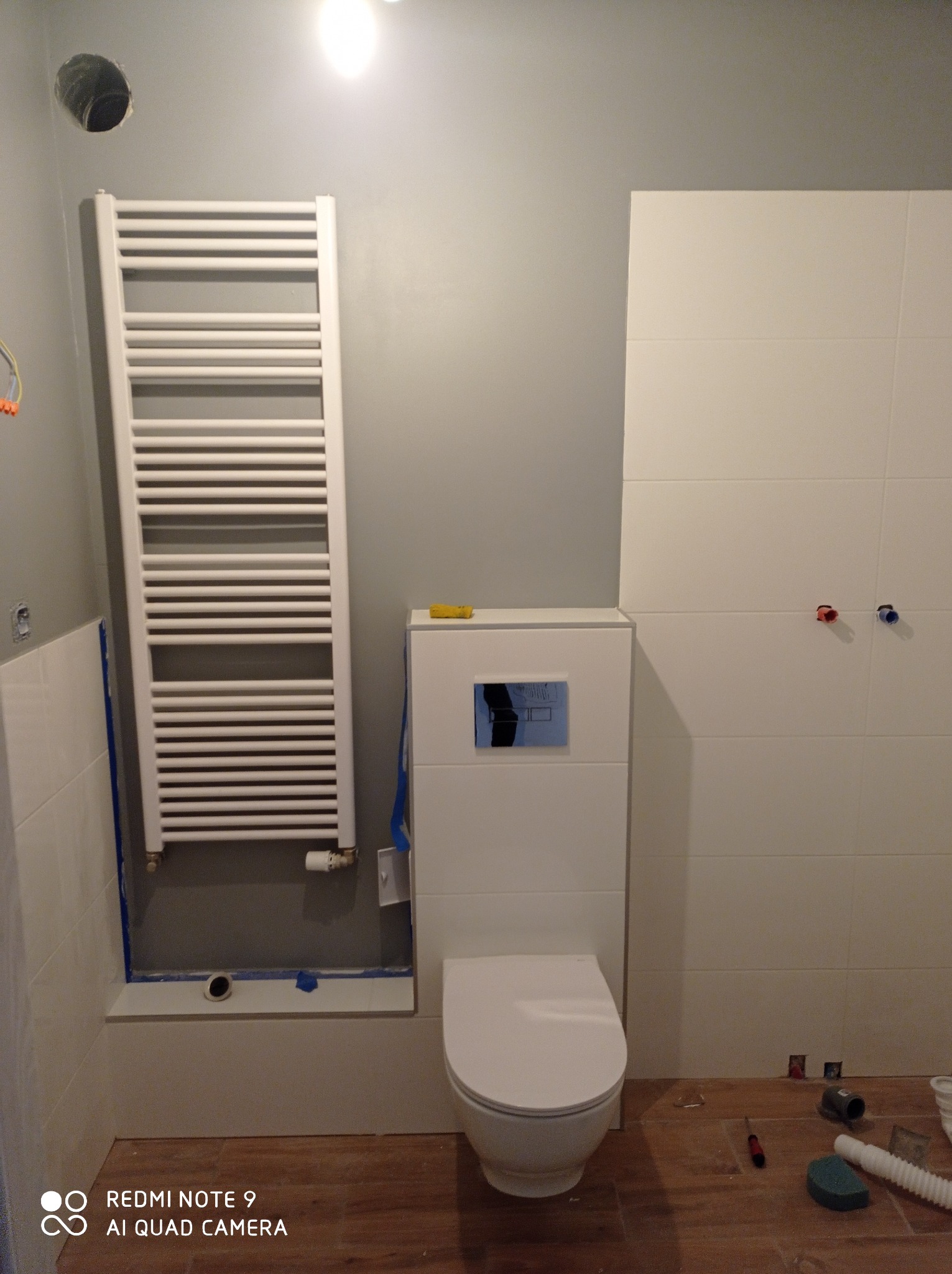 Wnętrze łazienki w trakcie remontu: biała toaleta z zabudowaną spłuczką, grzejnik drabinkowy, częściowo położone białe płytki, widoczne instalacje wodne i narzędzia na podłodze z paneli.