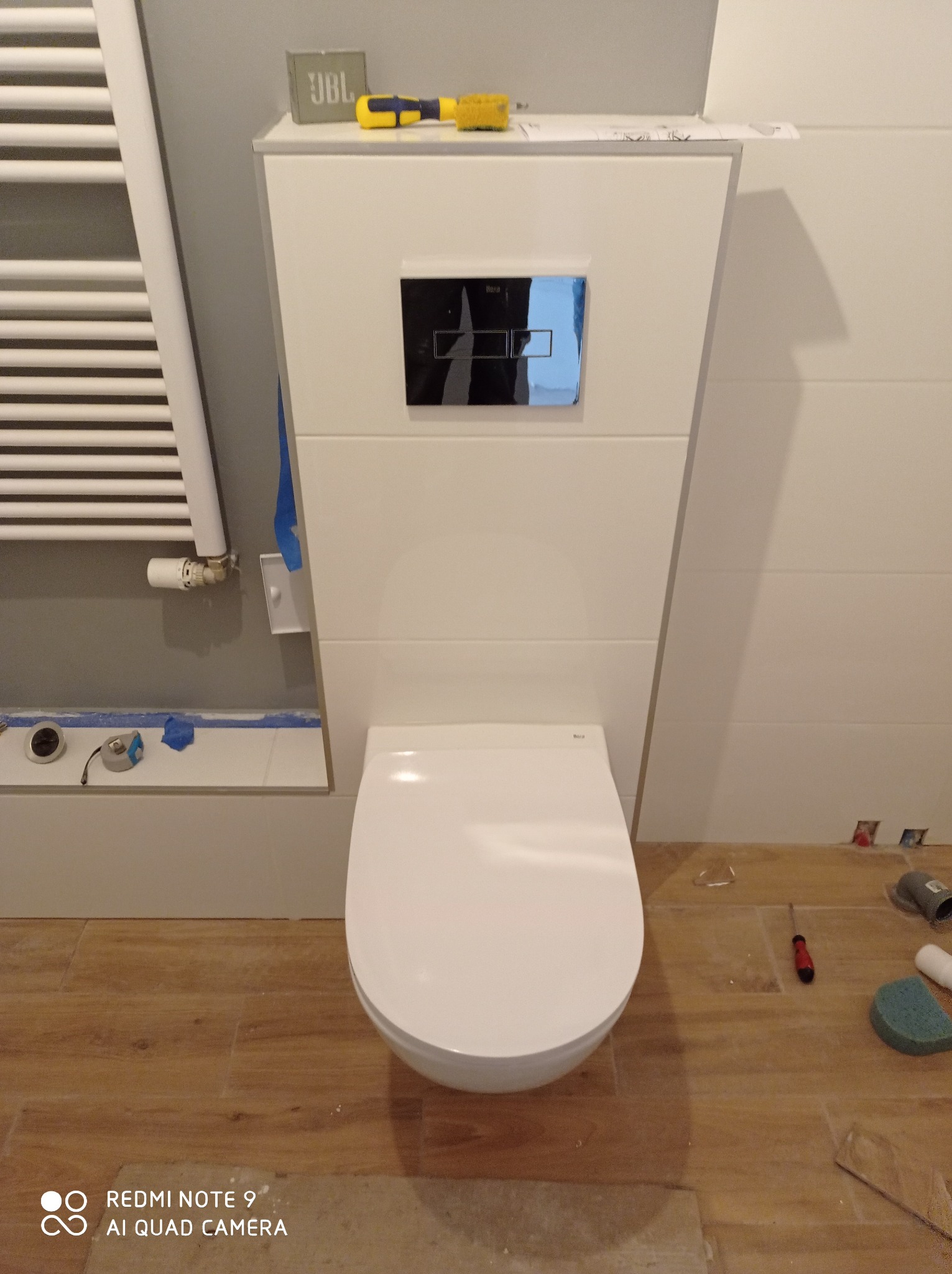 Podwieszana toaleta z białą zabudową i czarnym panelem spłukującym, widoczne narzędzia i akcesoria na podłodze z płytek imitujących drewno, grzejnik drabinkowy po lewej stronie, etap montażu.