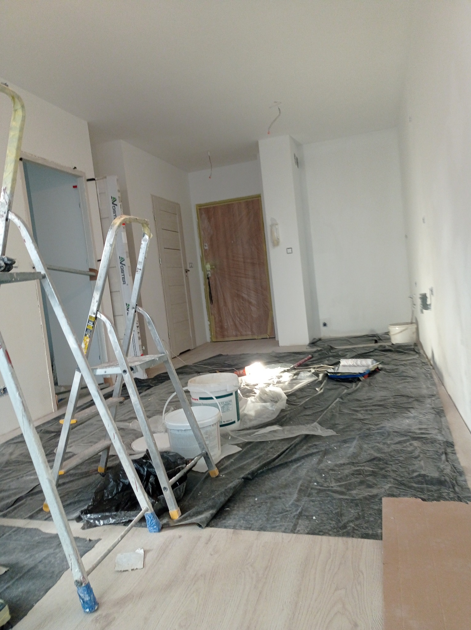 Pokój w trakcie odświeżania: zabezpieczona podłoga, drabiny malarskie, wiadra z farbą, wałek i kuweta malarska, białe ściany po malowaniu.