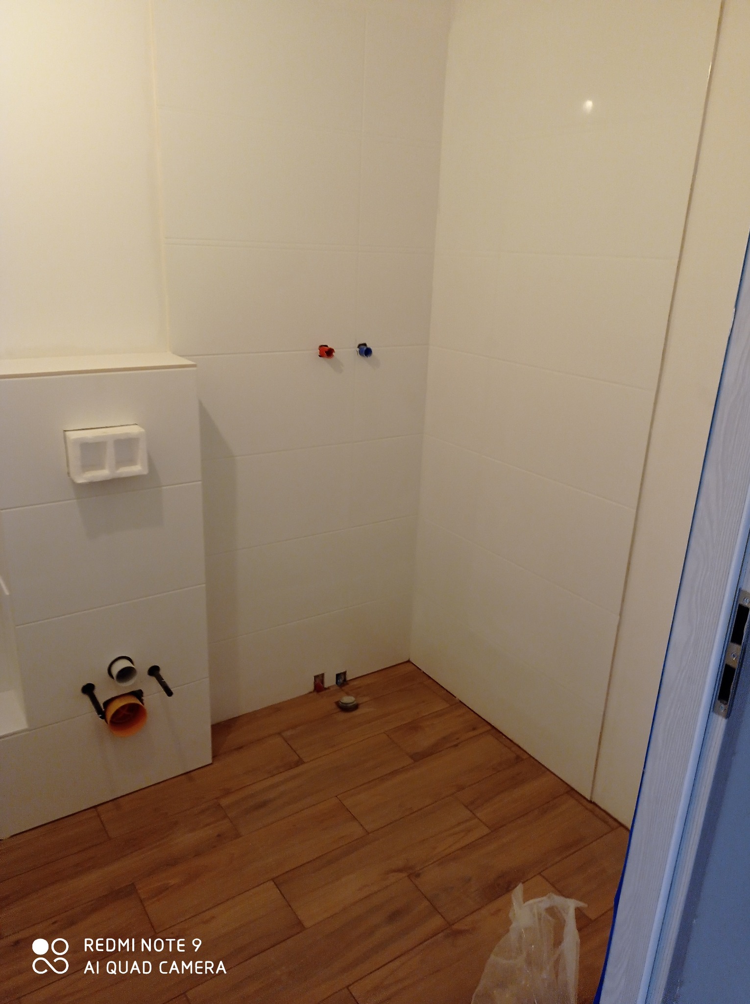 Łazienka w trakcie przygotowania do montażu armatury: białe płytki na ścianach, drewnopodobna podłoga, widoczne przyłącza wodne i odpływowe, częściowo zamontowany stelaż podtynkowy WC.