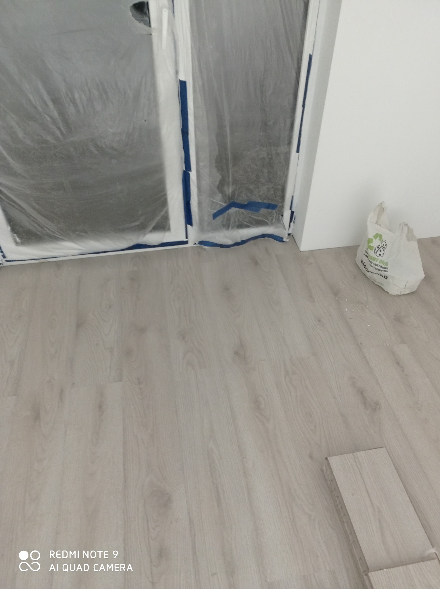 Podłoga z paneli w odcieniach szarości, zabezpieczone folią okna i biała ściana podczas prac wykończeniowych, widoczny worek z klejem