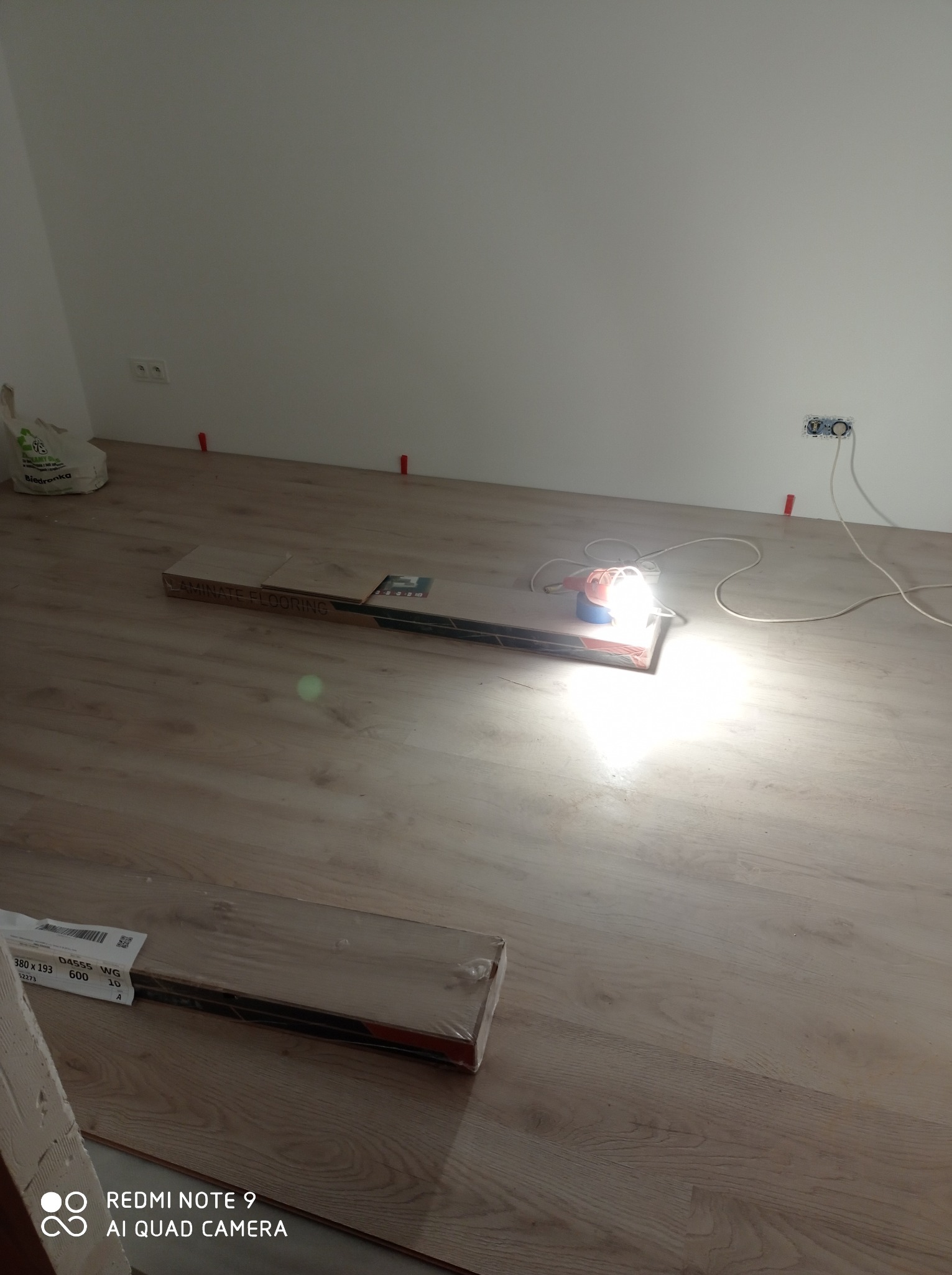 Układanie paneli podłogowych: paczki paneli, lampa budowlana oświetlająca fragment podłogi, dystanse przy ścianie, gniazdka elektryczne w tle.