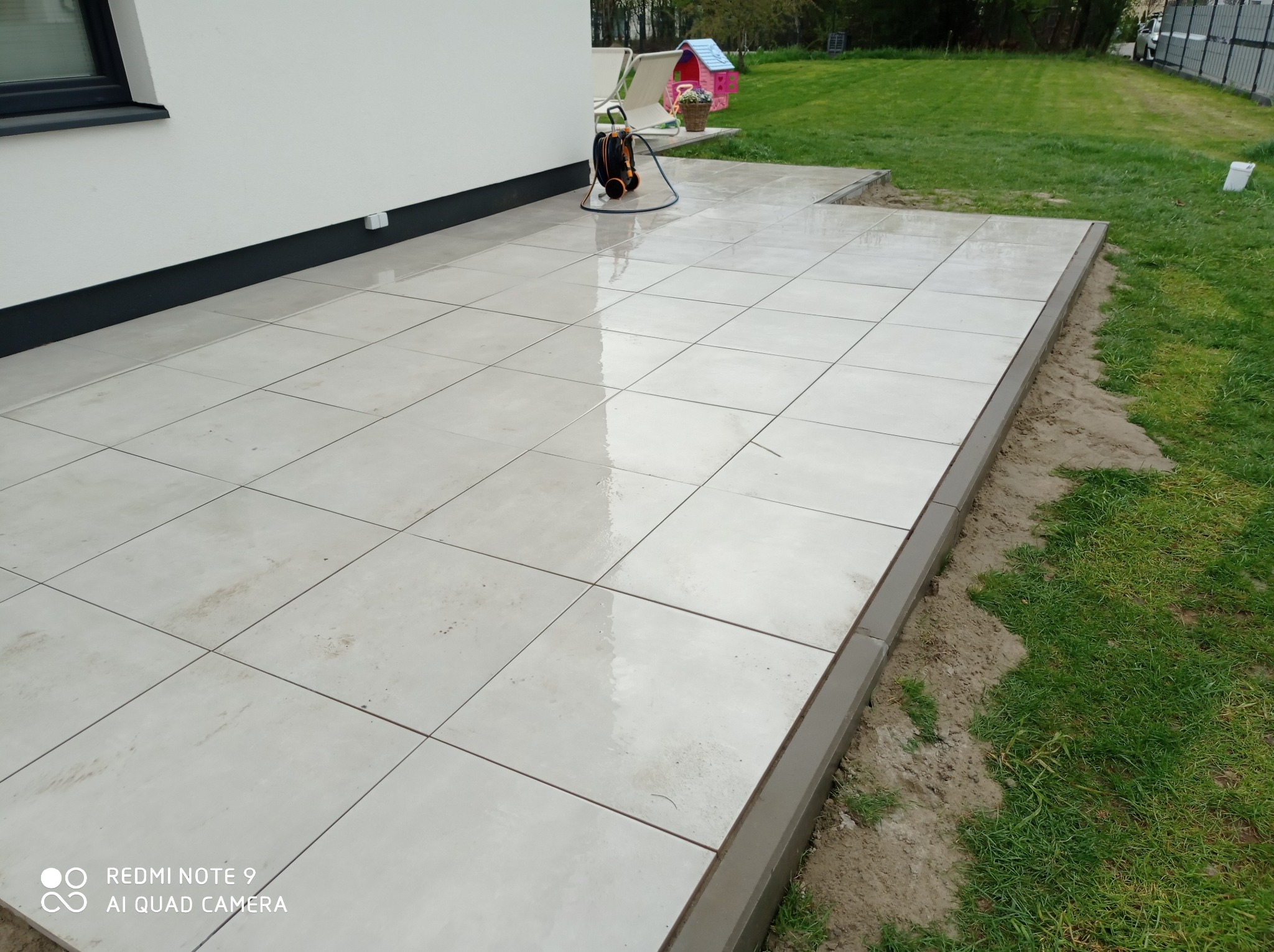 Jasne płytki tarasowe z szarą obwódką, świeżo po ułożeniu, z widoczną myjką ciśnieniową w tle, na tle zielonego trawnika i białego budynku.
