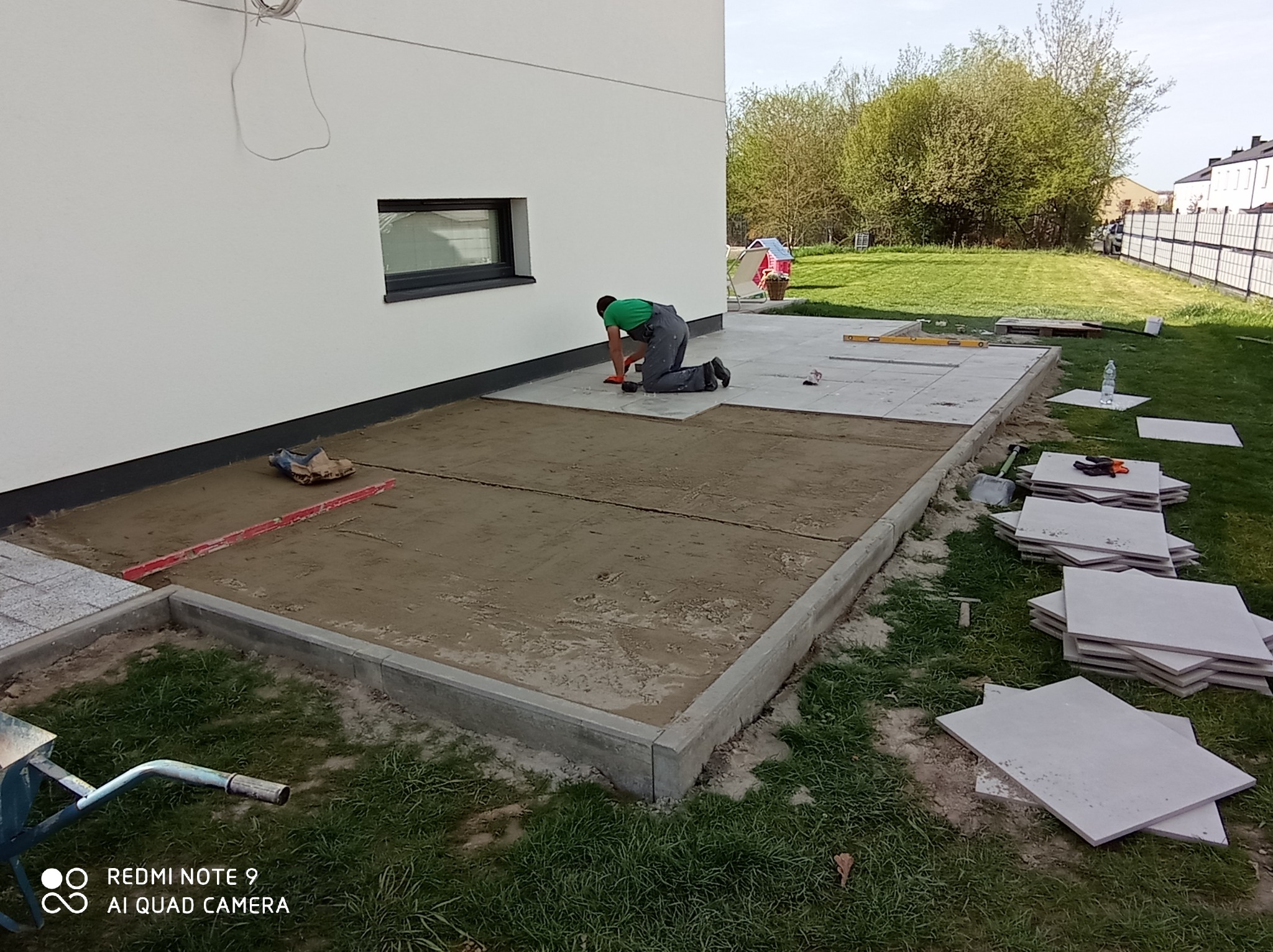 Układanie płyt tarasowych na zewnątrz budynku. Widoczny pracownik na kolanach, poziomica, płyty i przygotowane podłoże. Częściowo ułożona nawierzchnia.