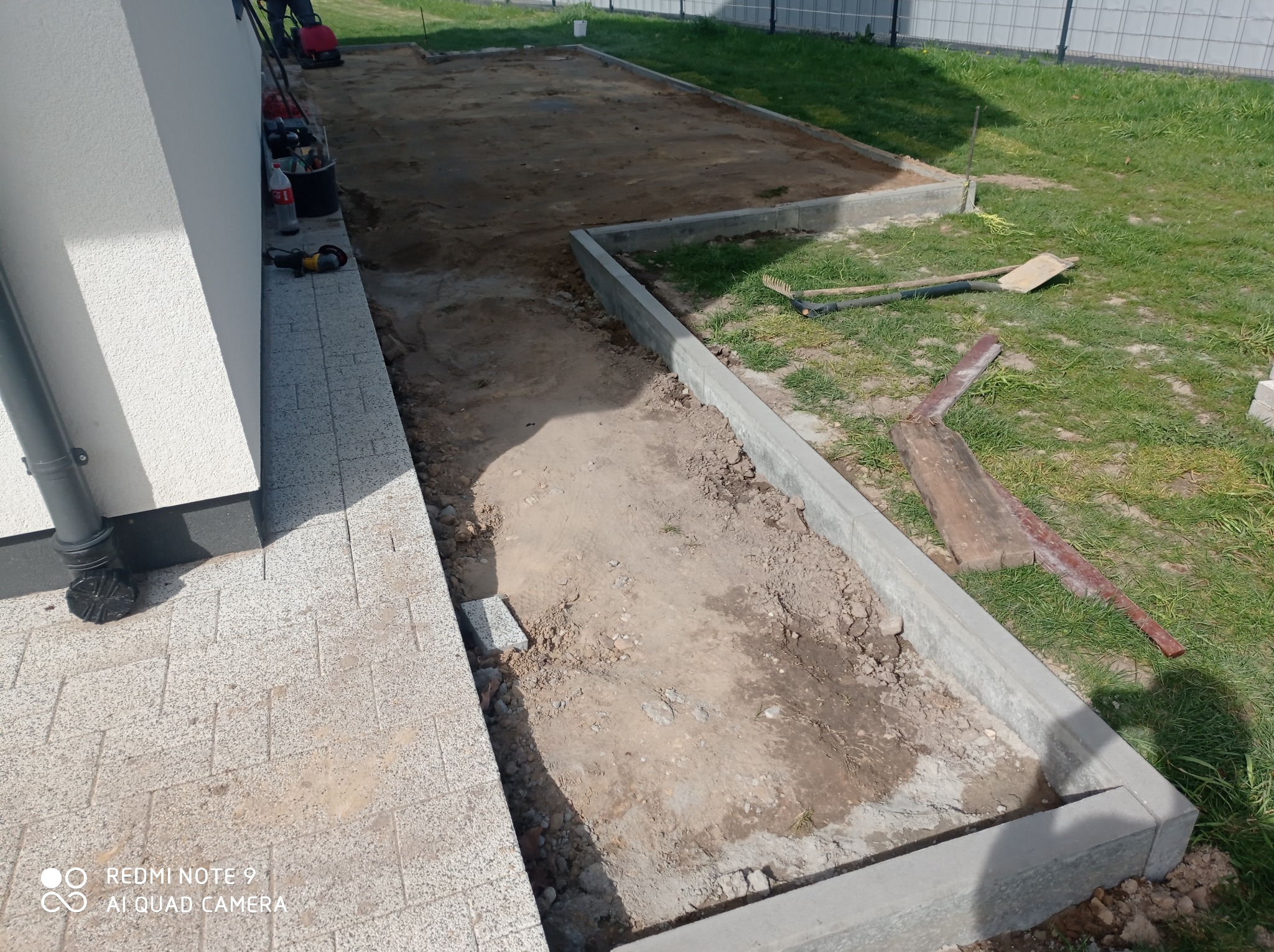 Przygotowanie terenu pod kostkę: obrzeża betonowe, ubita ziemia, narzędzia pracy obok budynku. Widok z góry, etap prac brukarskich.