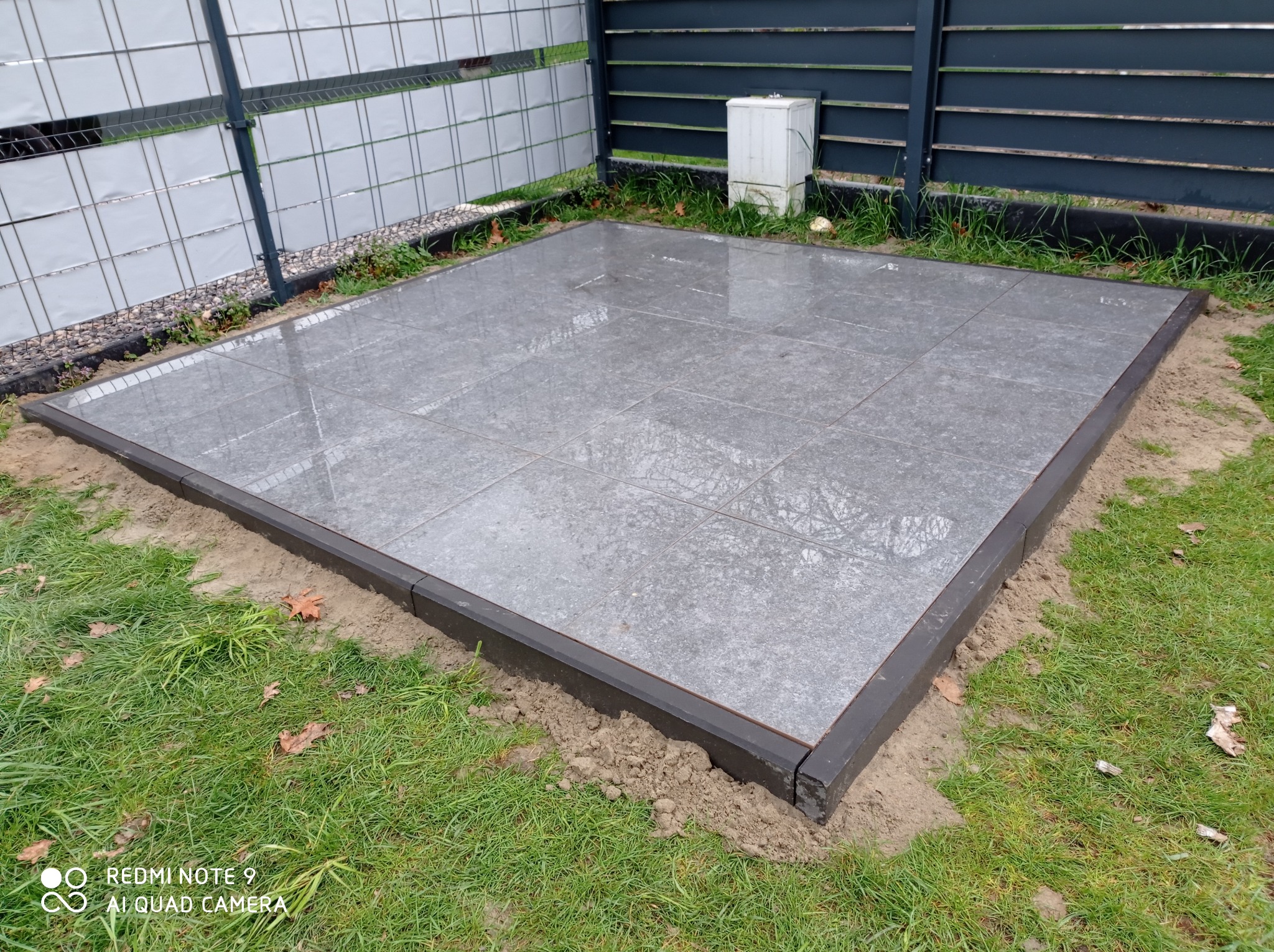 Wykończony taras z szarych płyt z ciemną obwódką, otoczony trawnikiem i ogrodzeniem. Widoczne odbicia na gładkiej powierzchni płyt.