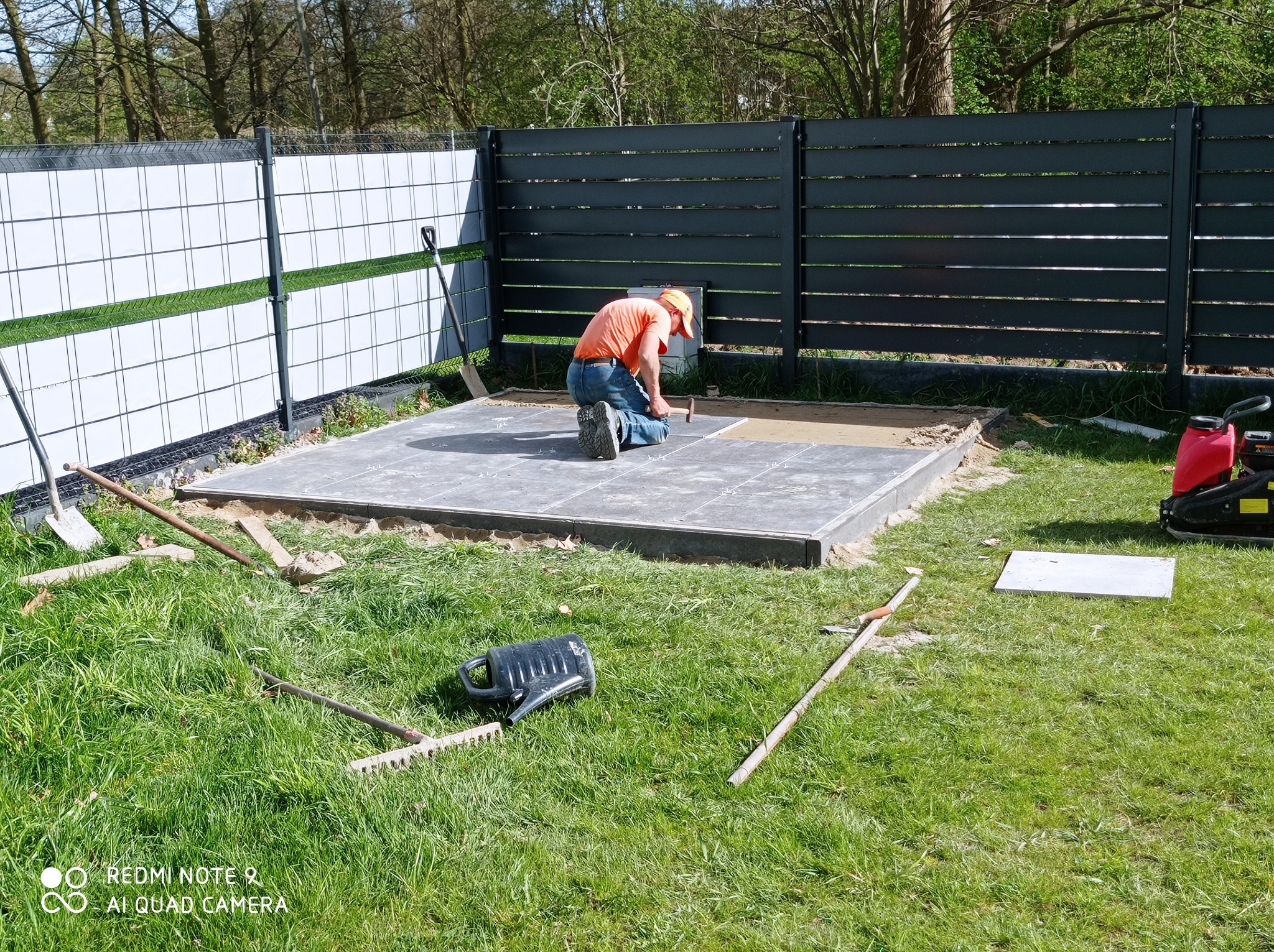 Pracownik w pomarańczowej koszulce klęczy, układając szare płyty chodnikowe na przygotowanym podłożu. W tle widoczne ogrodzenie i trawnik z narzędziami.