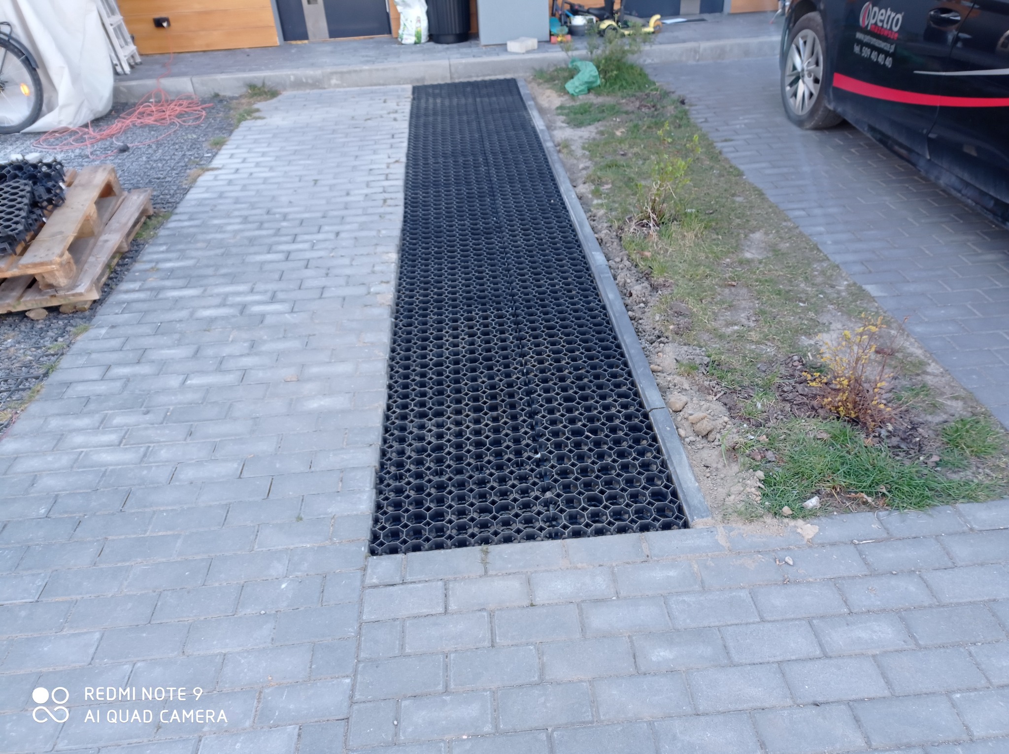 Szara kostka brukowa z liniowym odwodnieniem w postaci kratki. Częściowo widoczny samochód i paleta z elementami do układania kostki.