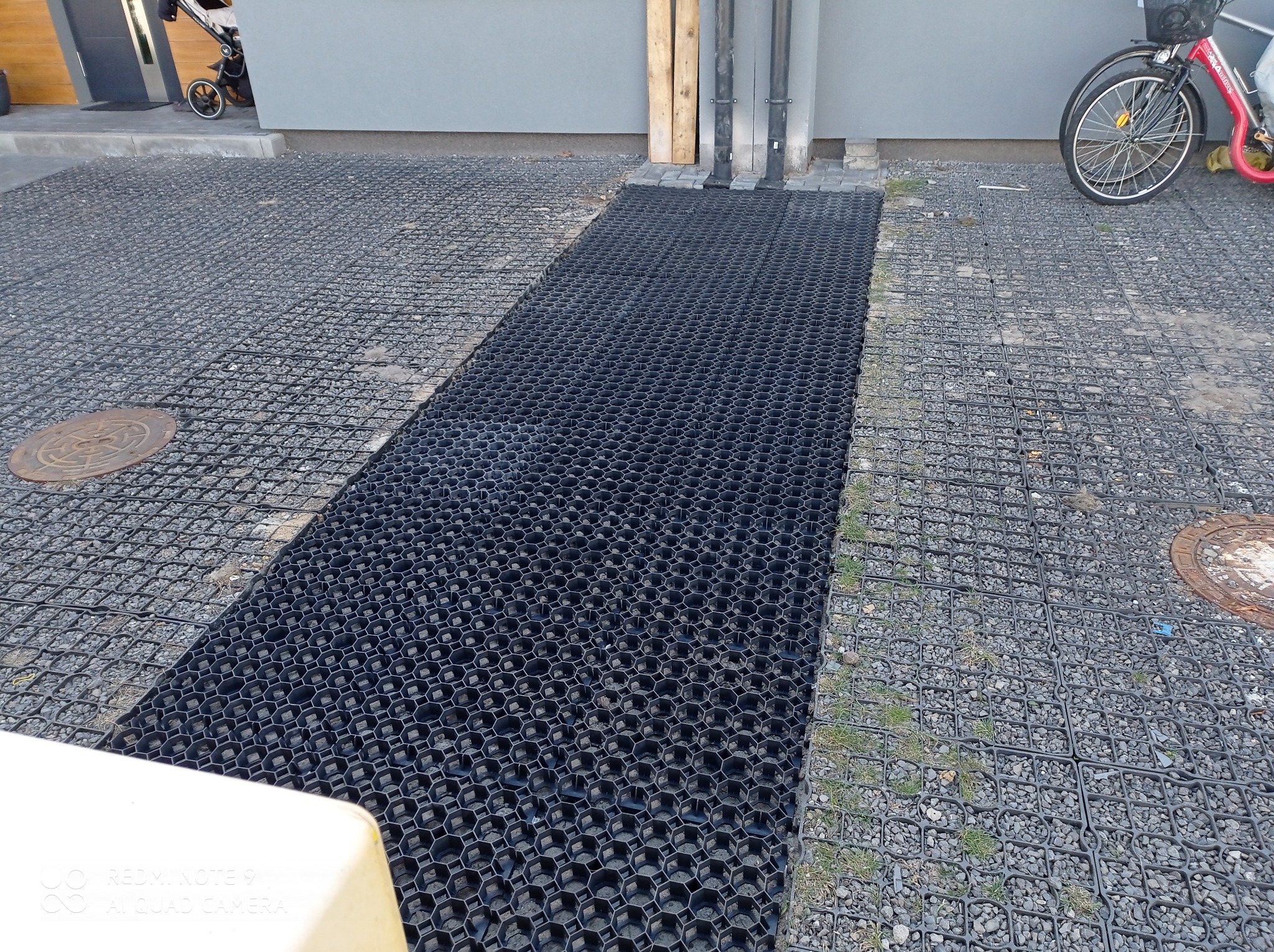 Nietypowe czarne kratki trawnikowe wkomponowane w szarą nawierzchnię z kamyczków. W tle widać wózek dziecięcy i rower, co sugeruje przestrzeń przed domem.