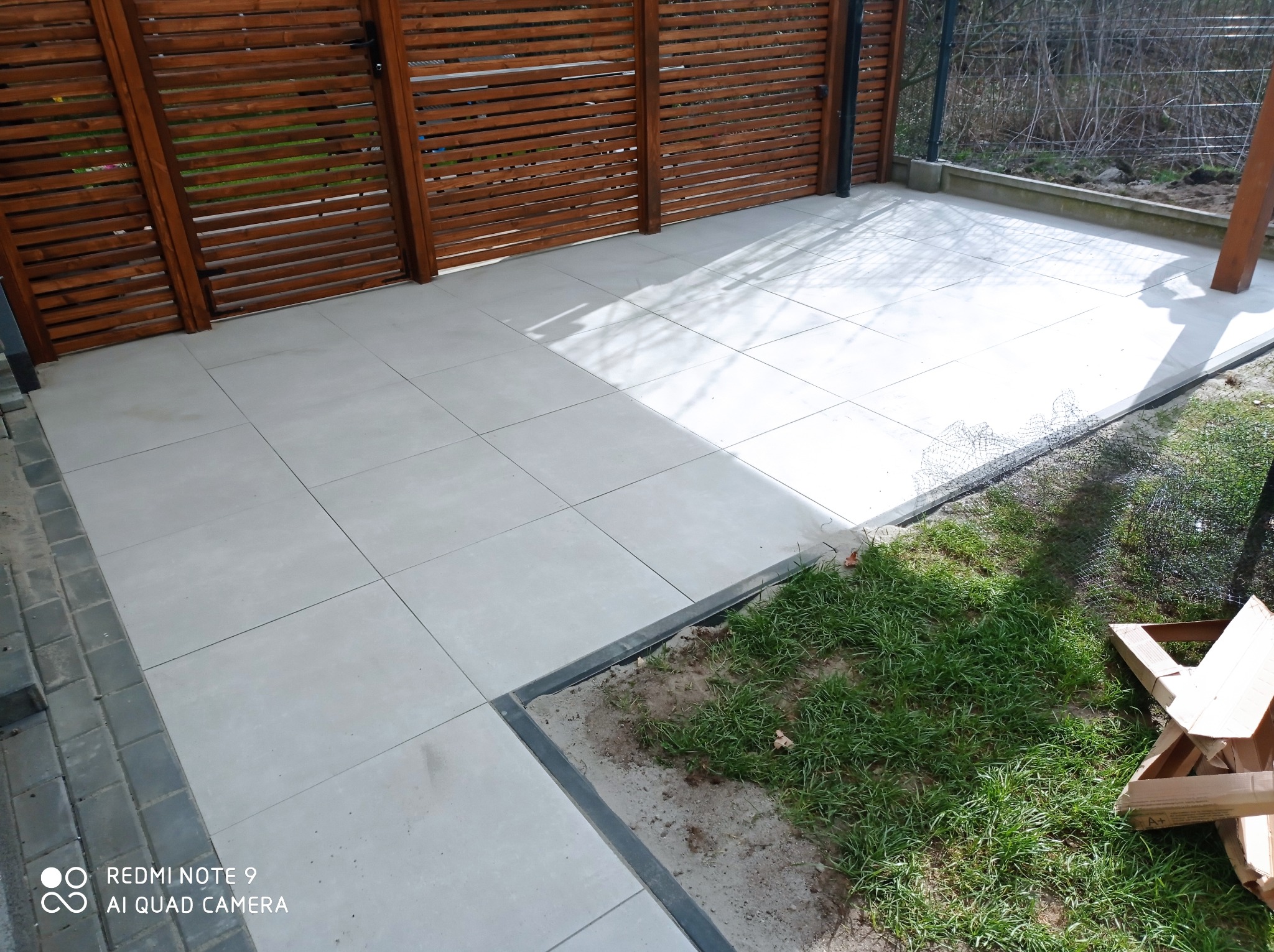 Jasnoszara, kwadratowa płytka tarasowa z ciemnoszarą obwódką, ułożona przy drewnianej konstrukcji w ogrodzie. Część trawnika widoczna w prawym dolnym rogu.