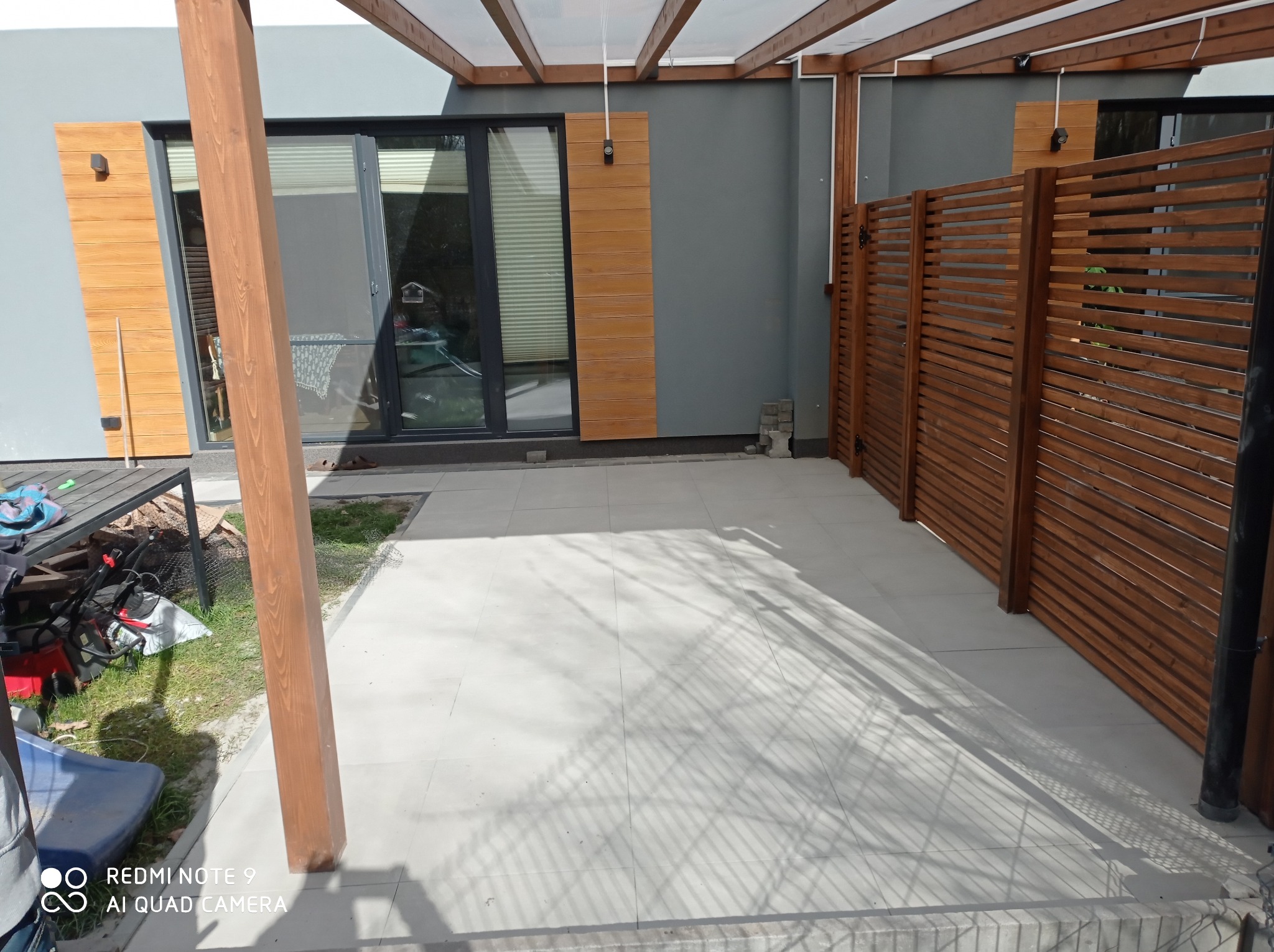 Wyłożony jasnymi płytami taras z drewnianą pergolą, przylegający do szarego budynku z drewnianymi elementami. Widoczny fragment drewnianego ażurowego ogrodzenia.