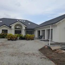 Biały dom jednorodzinny z ciemną dachówką i widocznymi śladami prac wykończeniowych elewacji, rusztowania i materiały budowlane na podjeździe z kostki brukowej, donice z kwiatami przy wejściu.