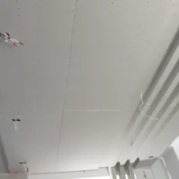 Montaż sufitu podwieszanego z płyt kartonowo-gipsowych, widoczne kable wystające z otworów na oświetlenie, oraz elementy konstrukcyjne z drewna.