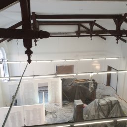 REMONTY DAMIAN DUSZCZAK - Wnętrze w trakcie prac wykończeniowych, widok z góry: montaż ścianek działowych z płyt kartonowo-gipsowych, zabezpieczone meble folią, widoczne drewniane elementy konstrukcyjne pod sufitem.