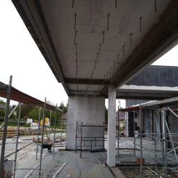 REMONTY DAMIAN DUSZCZAK - Wewnętrzne ujęcie surowego stanu budynku w trakcie budowy, widoczne betonowe stropy z wystającymi prętami, rusztowania i elementy konstrukcyjne.